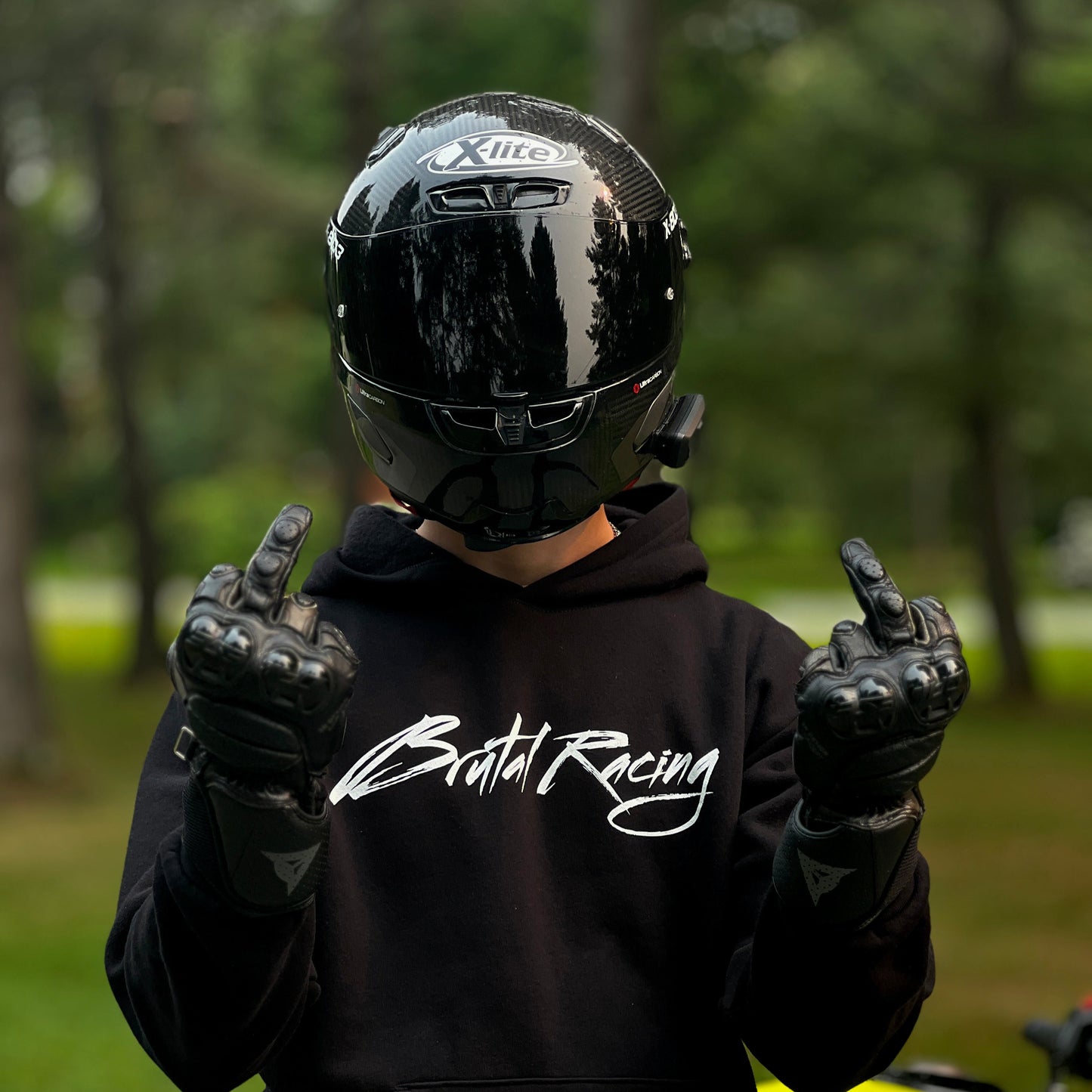 Person wearing a black helmet, gloves, and hoodie with 'Brutal Racing' text, standing next to a yellow motorcycle.